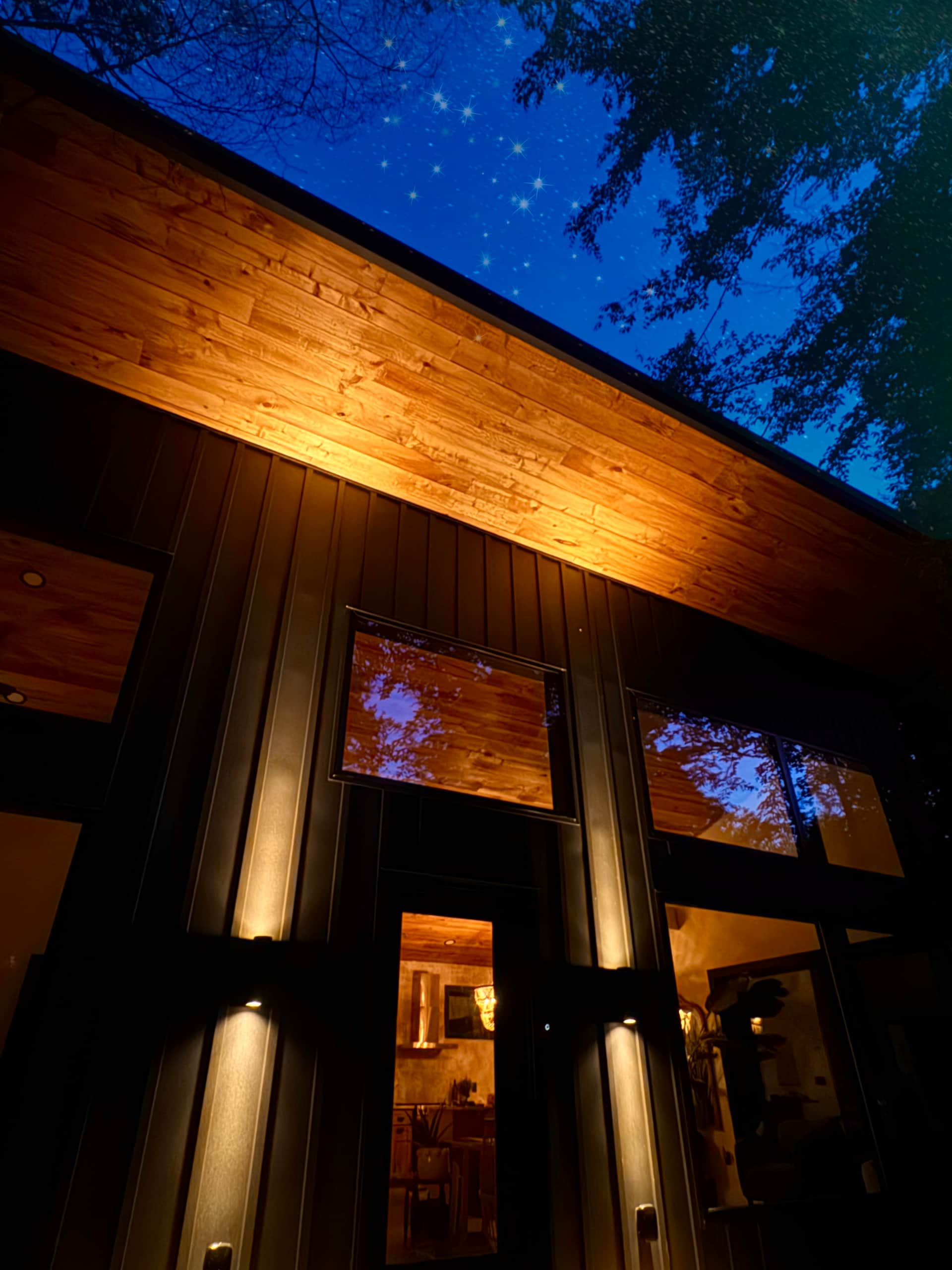 Warm interior shot of the cabin's main living area — floor-to-ceiling windows overlooking the forest, a cozy fireplace, warm wood tones and soft furnishings at golden hour